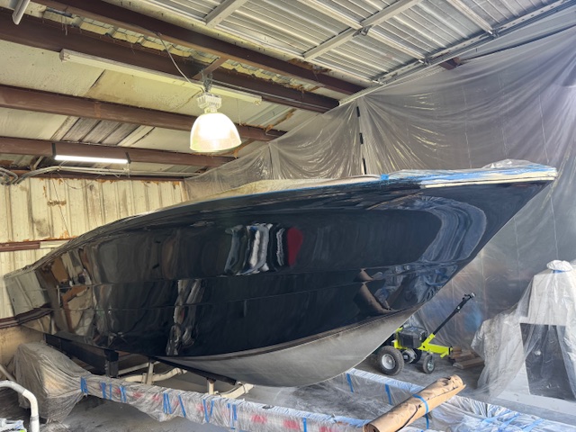 Freshly painted gloss black boat hull inside the Harper Marine Services shop, masked off with plastic sheeting after spraying.